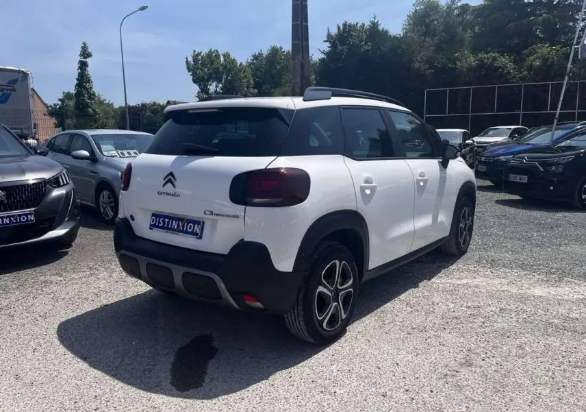 Vue 3/4 arrière droite d'un Citroën C3 Aircross blanc Banquise avec barres de toit noir brillant sur parking.