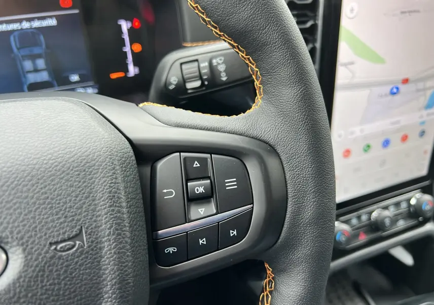 Gros plan sur le volant avec surpiqûres orange du Ford Ranger 2025 gris carbone, tableau de bord et écran tactile visibles.