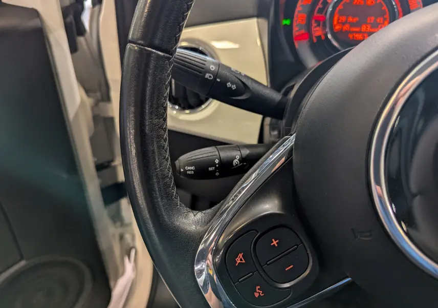 Gros plan sur le volant noir de la Fiat 500 blanc Bossanova, avec commandes audio et tableau de bord rouge en arrière-plan.