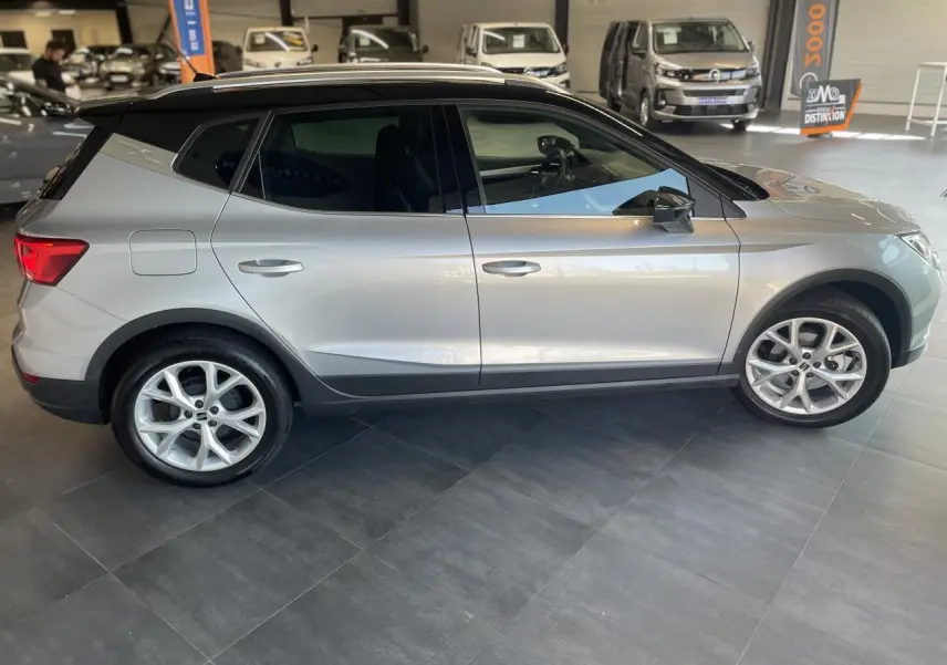 Vue latérale droite d'un SEAT Arona 2024 gris clair avec toit noir et jantes alliage 16 pouces en showroom.