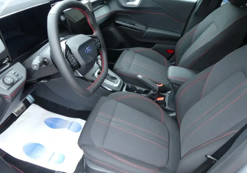 Vue intérieure côté conducteur du Ford Puma 2025, sièges tissu gris avec surpiqûres rouges et volant sport cuir.