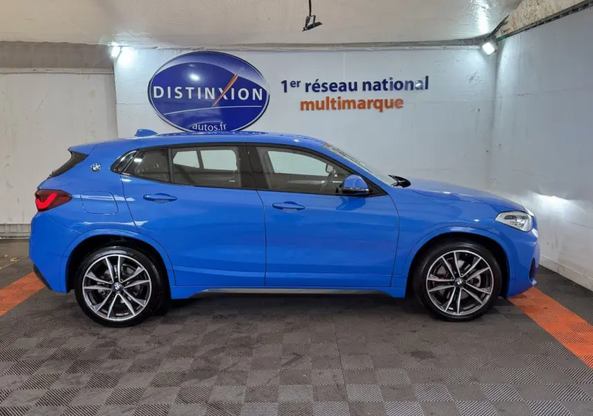 Vue de profil côté gauche d’un BMW X2 xDrive25e M Sport bleu dans un garage avec logo Distinxion en arrière-plan.