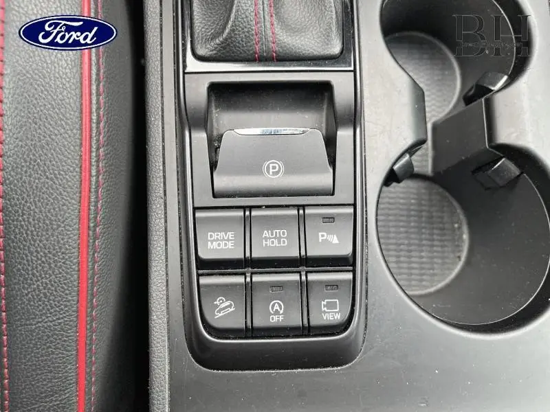 Gros plan sur la console centrale noire avec boutons de contrôle et surpiqûres rouges visibles sur le cuir adjacent