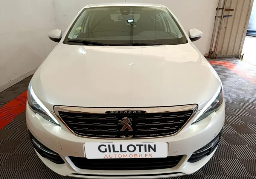 Vue avant frontale d'une Peugeot 308 blanche avec calandre chromée et feux LED allumés dans un garage.