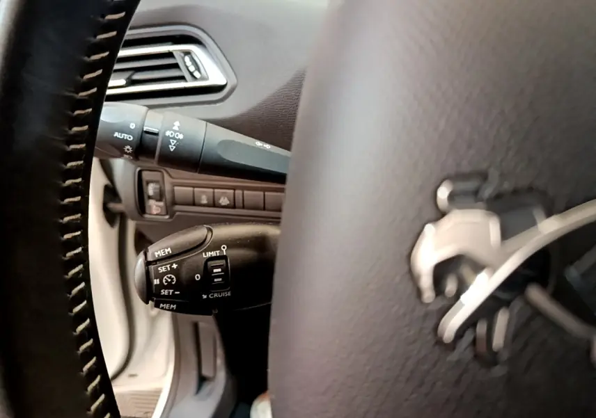Gros plan sur le volant cuir noir de la Peugeot 308 blanc, avec commandes de régulateur de vitesse visibles à gauche.