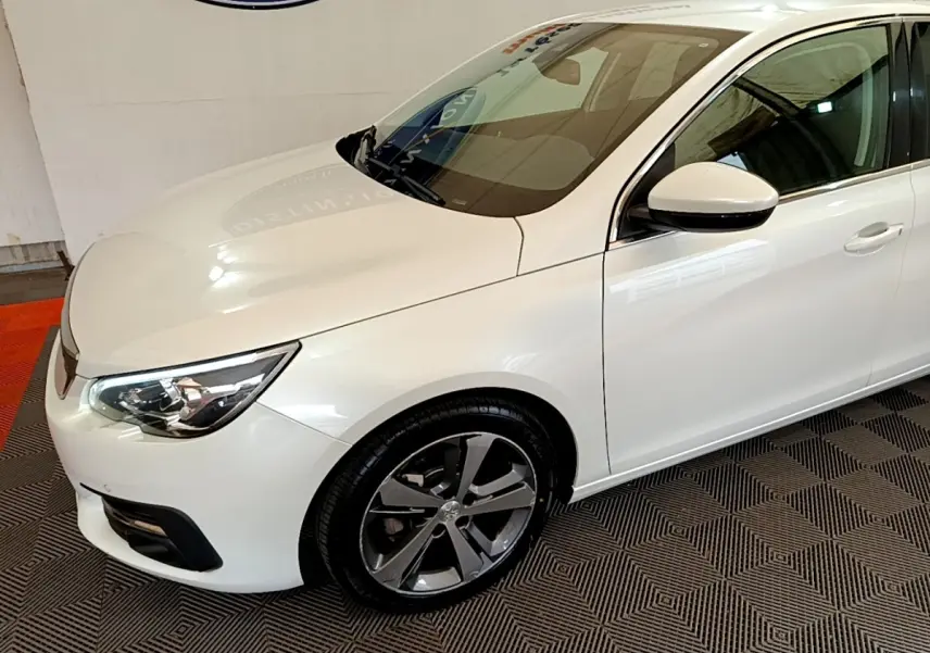 Vue 3/4 avant droit d'une Peugeot 308 blanche avec jantes alliage et phares LED ellipsoïdaux visibles.