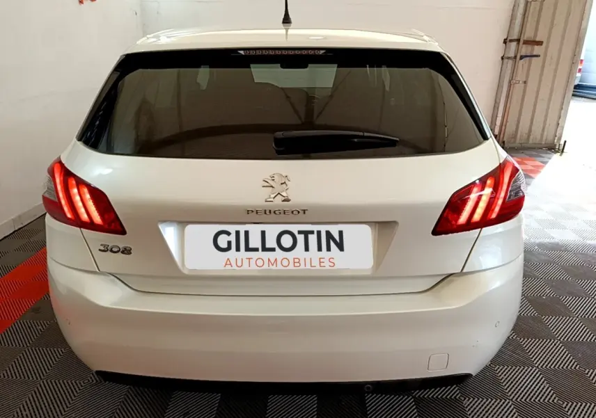 Vue arrière d'une Peugeot 308 blanche de 2017 avec feux arrière allumés et logo visible sur un sol carrelé.