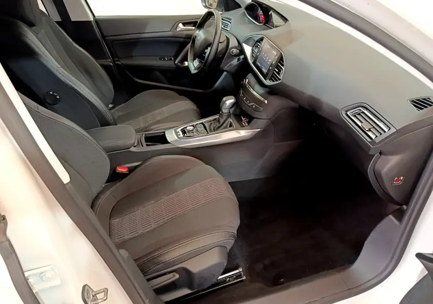 Vue intérieure côté conducteur de la Peugeot 308 blanche, montrant sièges noirs et tableau de bord avec écran tactile.
