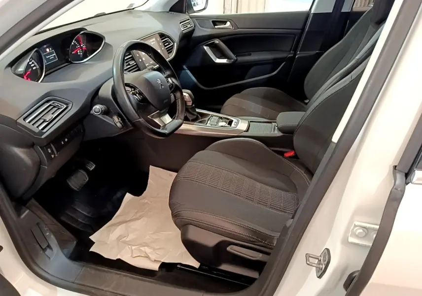 Vue intérieure côté conducteur de la Peugeot 308 blanche, montrant sièges noirs et tableau de bord avec écran tactile.