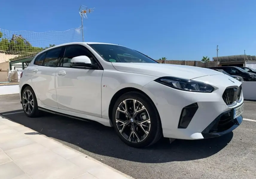 BMW Série 1 blanche vue 3/4 avant droit, avec jantes alliage bicolores et calandre M Sport sous un ciel bleu.