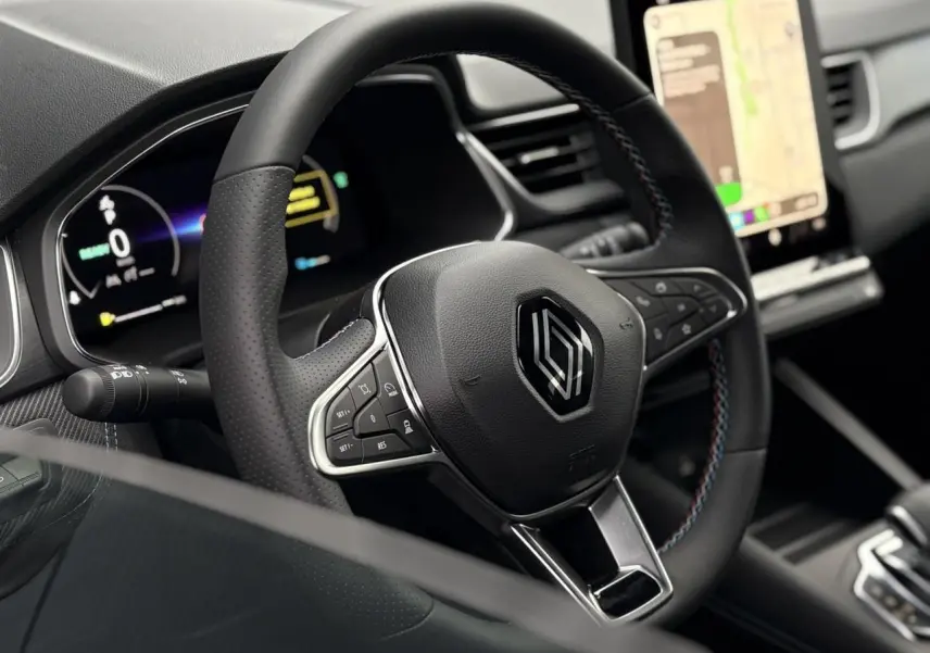 Vue rapprochée du volant noir avec logo Renault et surpiqûres tricolores, tableau de bord numérique et écran tactile visibles.