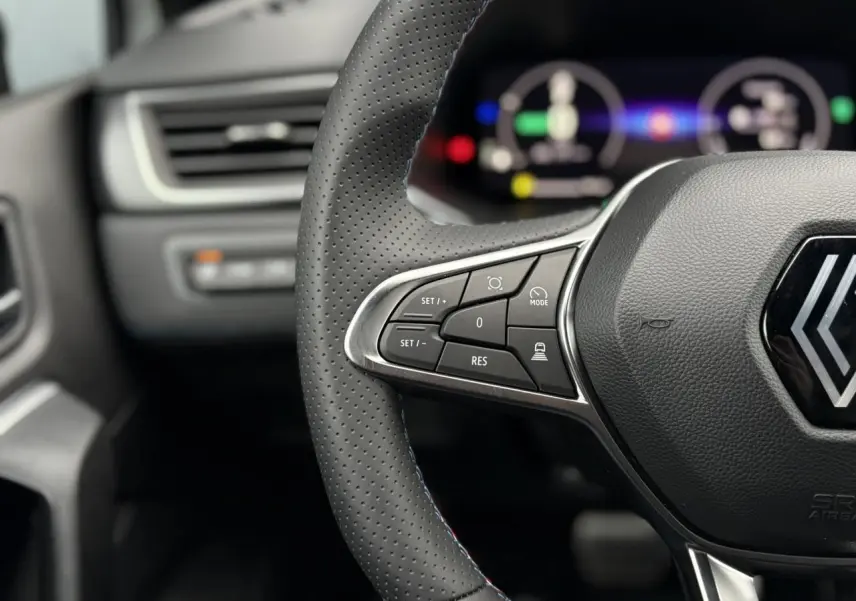 Gros plan sur le volant perforé avec commandes intégrées et logo Alpine du Renault Captur Blanc Nacré, tableau de bord numérique flou en arrière-plan.