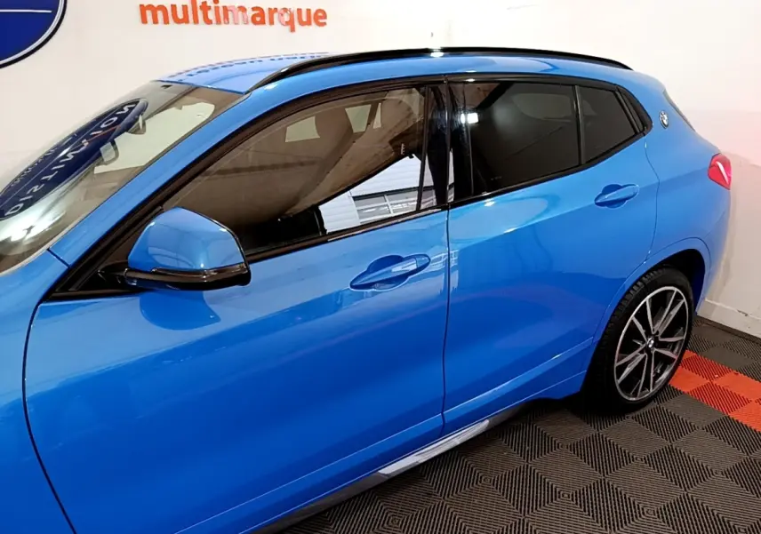 Vue côté gauche d'un BMW X2 sDrive20i M Sport bleu, avec jantes alliage et vitres teintées en intérieur showroom.