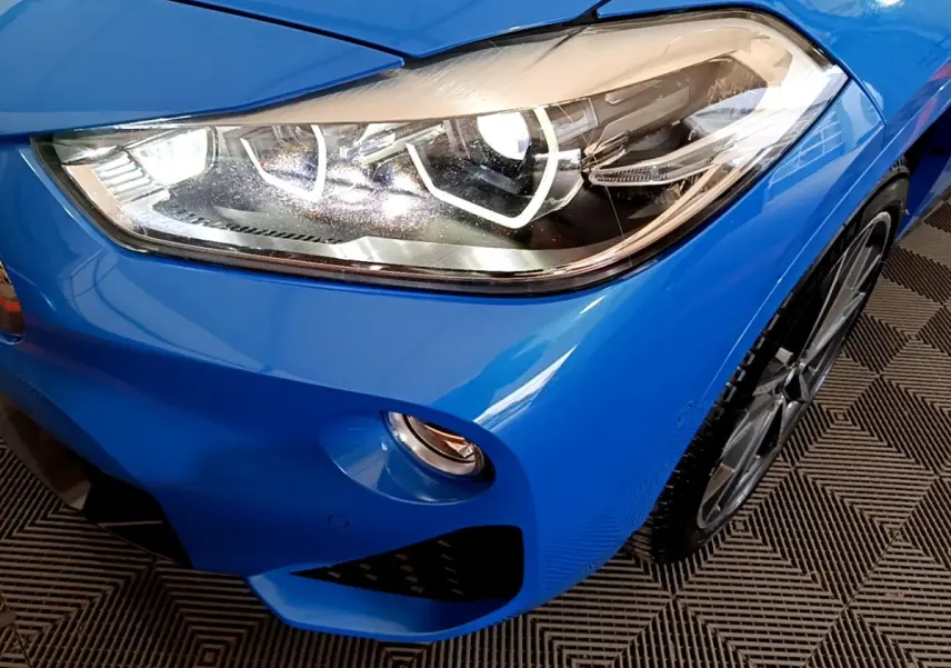 Gros plan sur le phare avant droit bleu vif d'une BMW X2 sDrive20i M Sport avec jante alliage visible.