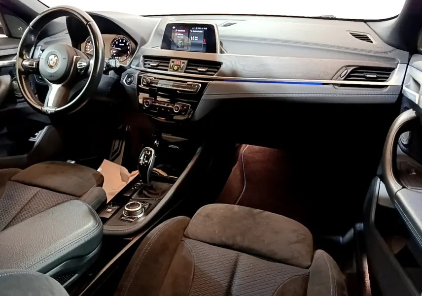 Intérieur BMW X2 sDrive20i M Sport 2019, vue avant côté conducteur, tableau de bord avec écran tactile et éclairage d’ambiance bleu.