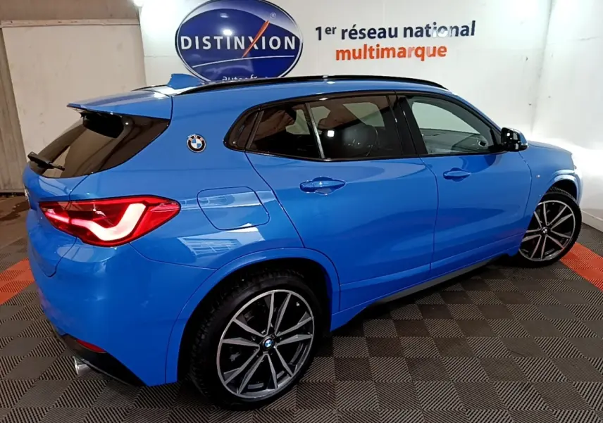 BMW X2 sDrive20i M Sport bleu en vue 3/4 arrière droit, avec jantes alliage et feux arrière allumés.