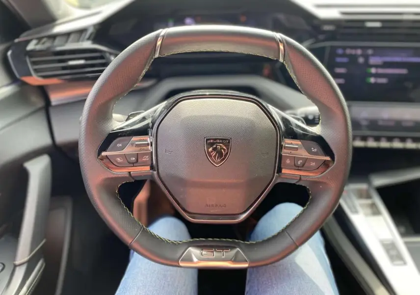 Volant cuir GT de la Peugeot 308 2023 vu de face, avec commandes intégrées et tableau de bord numérique flou.