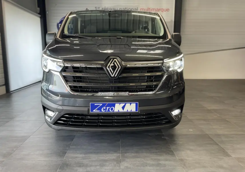 Vue frontale du Renault Trafic Fourgon gris Comète avec phares et feux antibrouillard allumés en intérieur showroom.