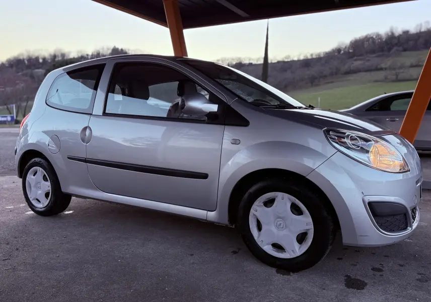Renault Twingo II gris vue de profil côté gauche sous abri, phare avant droit allumé, jantes avec enjoliveurs blancs.