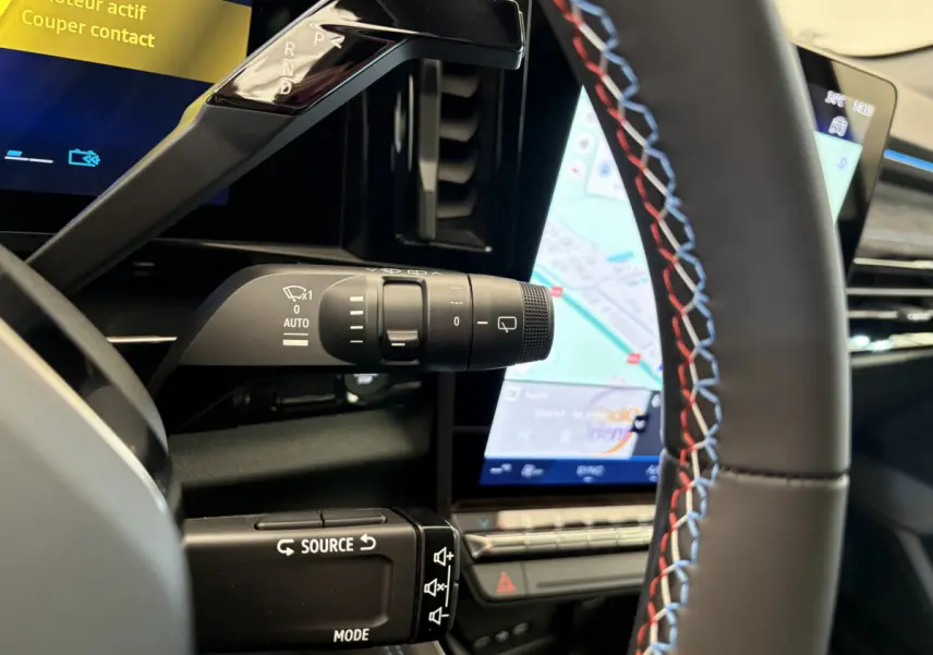 Gros plan intérieur sur le volant avec surpiqûres tricolores et commande multifonctions, écran tactile de navigation visible.