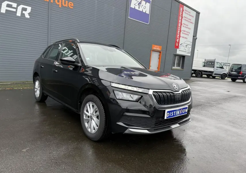 Vue 3/4 avant gauche d'un Skoda Kamiq noir magic nacré avec calandre chromée, stationné devant un bâtiment gris.