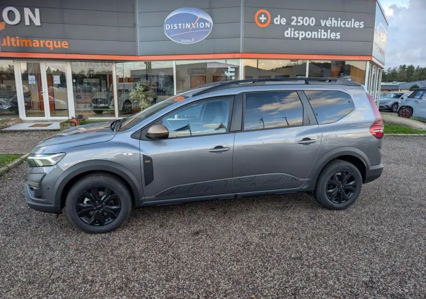 Vue de profil côté gauche d’un Dacia Jogger gris métallisé avec jantes noires et rétroviseurs brun cuivré, stationné devant un concessionnaire.