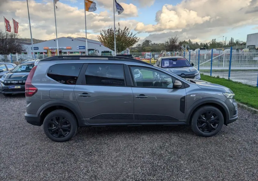 Vue de profil côté gauche d'un Dacia Jogger gris métallisé avec jantes noires et barres de toit noires.