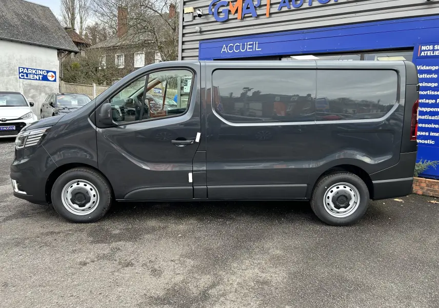 Renault Trafic Fourgon gris Comète vu de profil côté gauche, stationné devant un bâtiment commercial.