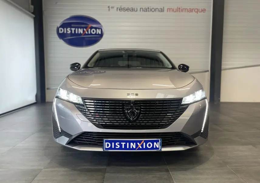 Vue frontale d'une Peugeot 308 gris Artense avec calandre noire et feux LED allumés en intérieur showroom.