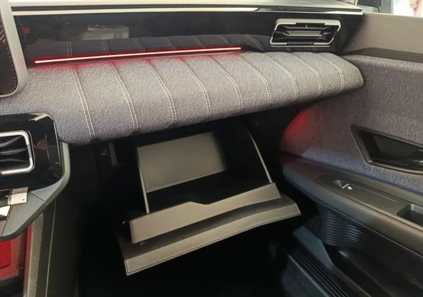 Intérieur de la Renault R5 Techno 150 ch 2025, vue côté passager sur la boîte à gants ouverte et tissu gris avec éclairage rouge.