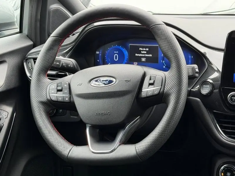 Vue rapprochée du volant sport perforé noir avec surpiqûres rouges du Ford Puma 2024, tableau de bord digital bleu en arrière-plan.