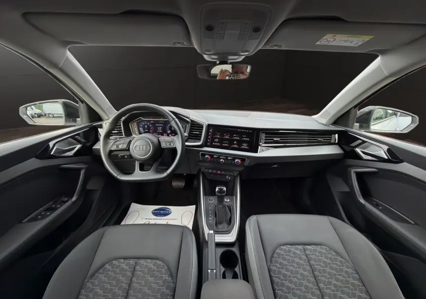 Vue intérieure avant de l'Audi A1 Sportback 2025 avec tableau de bord noir, volant sport cuir et écran tactile central.