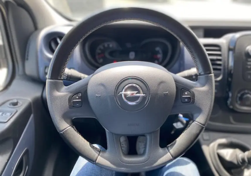 Vue rapprochée du volant noir de l'Opel Vivaro 2018 avec commandes intégrées et tableau de bord flou en arrière-plan.
