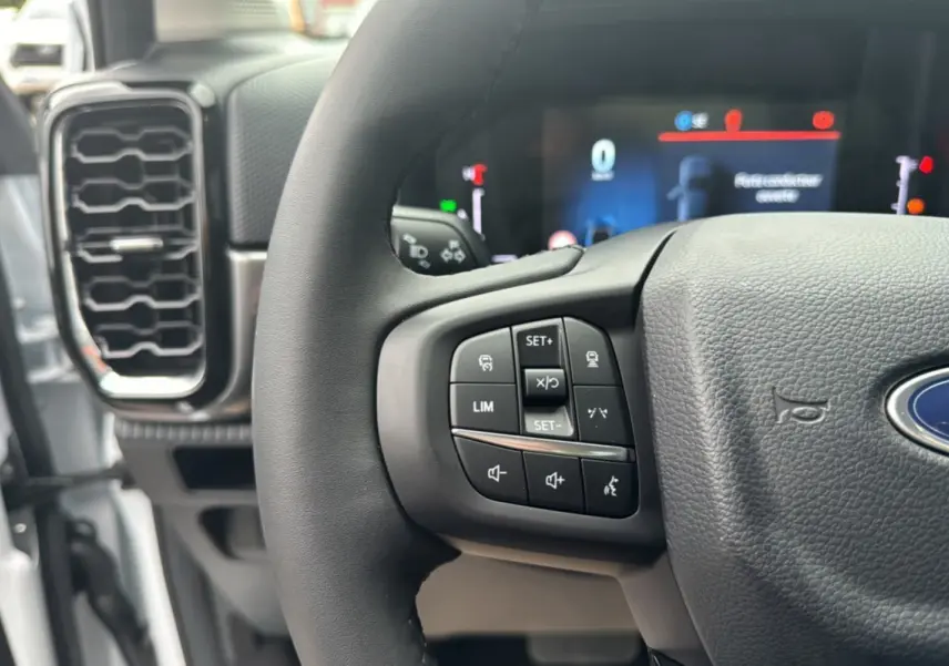 Gros plan sur le volant cuir multifonctions du Ford Ranger blanc glacier, avec commandes intégrées et tableau de bord numérique flou.