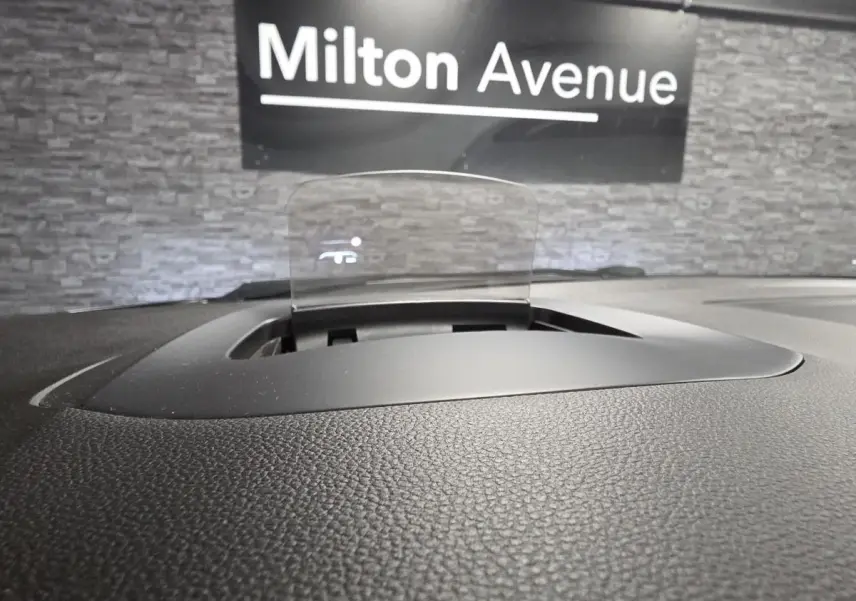 Affichage tête haute transparent sur le tableau de bord noir du Renault Scenic blanc, avec fond mural gris et panneau Milton Avenue.