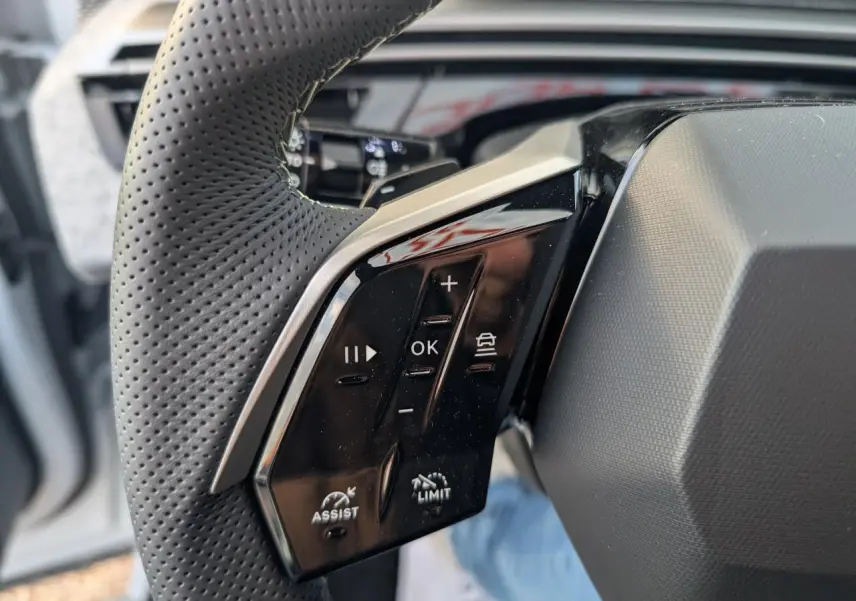 Gros plan sur les commandes noires à gauche du volant perforé gris du Peugeot 3008 blanc, version GT 2025.
