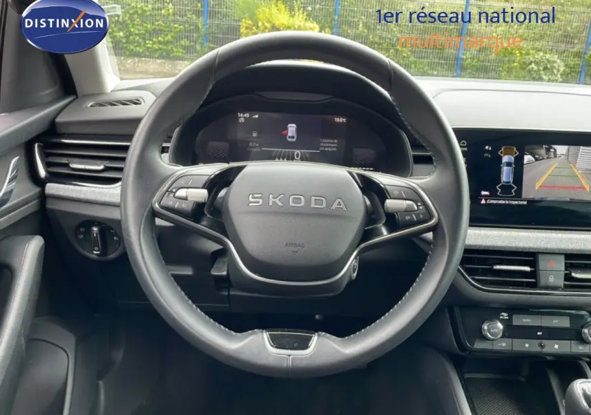 Vue intérieure centrée sur le volant SKODA noir avec tableau de bord numérique et écran de caméra de recul.