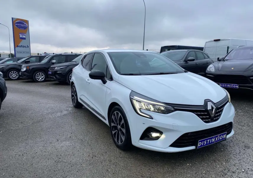 Renault Clio blanche vue 3/4 avant droit avec jantes alliage noires et feux LED allumés sur parking.