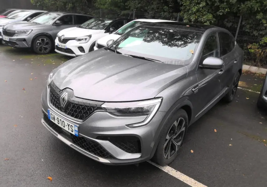 Renault Arkana gris métal foncé vu en 3/4 avant droit sur un parking sous un ciel couvert.