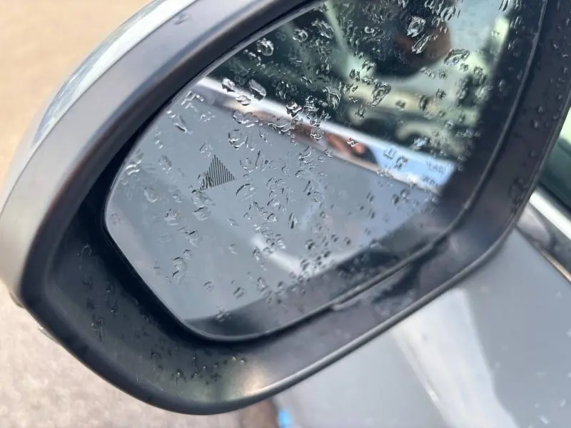 Rétroviseur extérieur côté gauche d'un Nissan Qashqai gris Argile 2024 avec gouttes d'eau et signal d'angle mort.