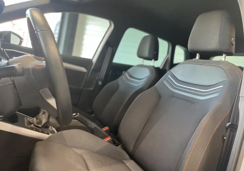 Vue intérieure côté conducteur du SEAT Arona 2024, sièges textile noirs avec surpiqûres orange et volant cuir multifonction.