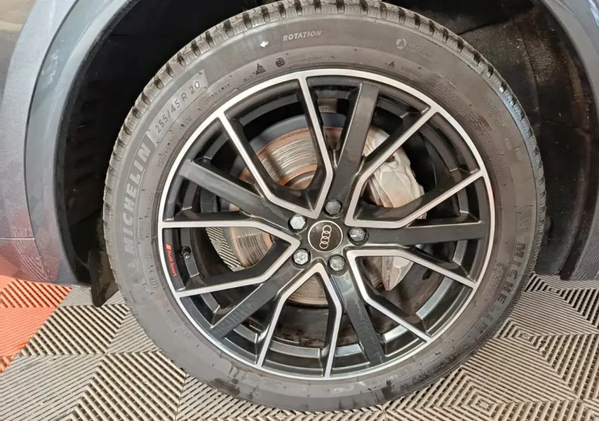 Gros plan sur la roue avant droite d’un Audi Q5 gris clair 2023, jante noire à détails argentés avec logo Audi au centre.
