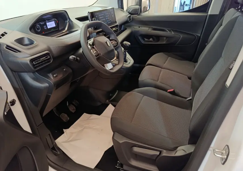 Vue intérieure côté conducteur du Peugeot Partner 2025 blanc, montrant tableau de bord, volant et sièges tissu gris.