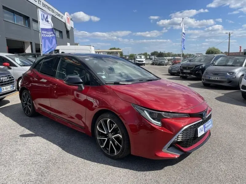 Toyota Corolla 184 H Collection rouge métal avec toit noir, vue 3/4 avant côté gauche sur parking extérieur.