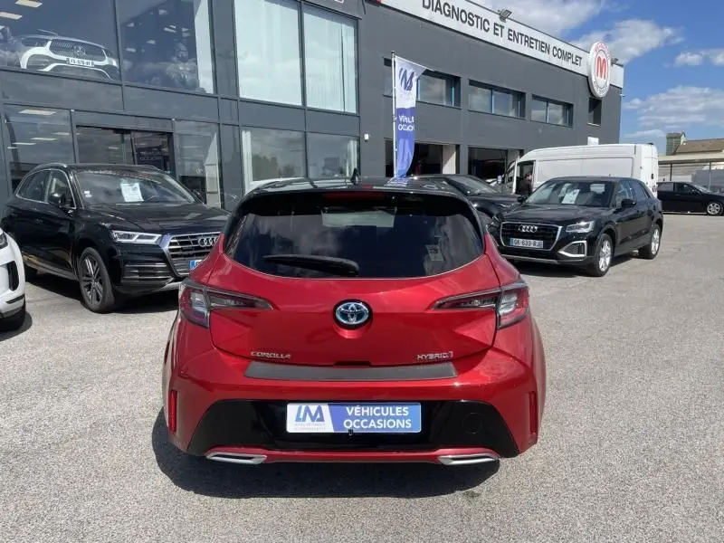Vue arrière d'une Toyota Corolla 184 H Collection rouge métal avec toit noir, stationnée devant un garage.