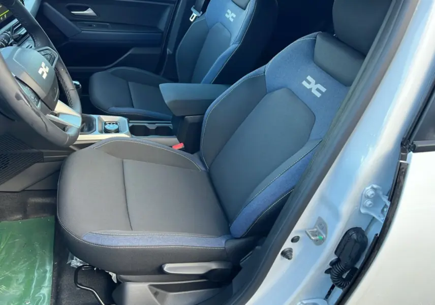 Vue intérieure côté conducteur du Dacia Duster blanc glacier 2025, sièges tissu bleu et noir avec logo distinctif brodé.