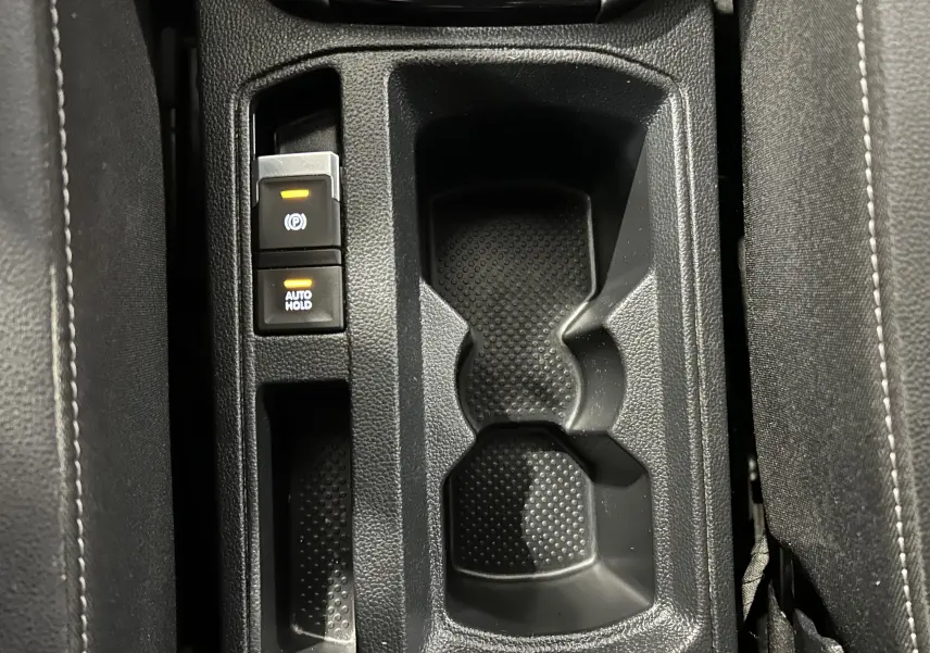 Vue en plongée sur la console centrale noire du Volkswagen T-Roc avec porte-gobelets et boutons frein à main et auto hold.