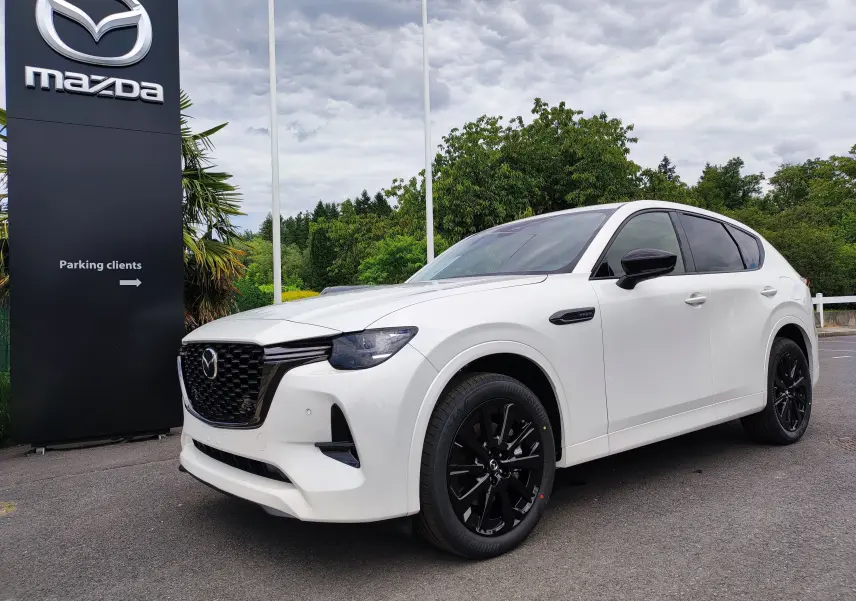 Vue 3/4 avant droit d'un Mazda CX-60 blanc métal avec jantes noires, devant un panneau Mazda en extérieur.
