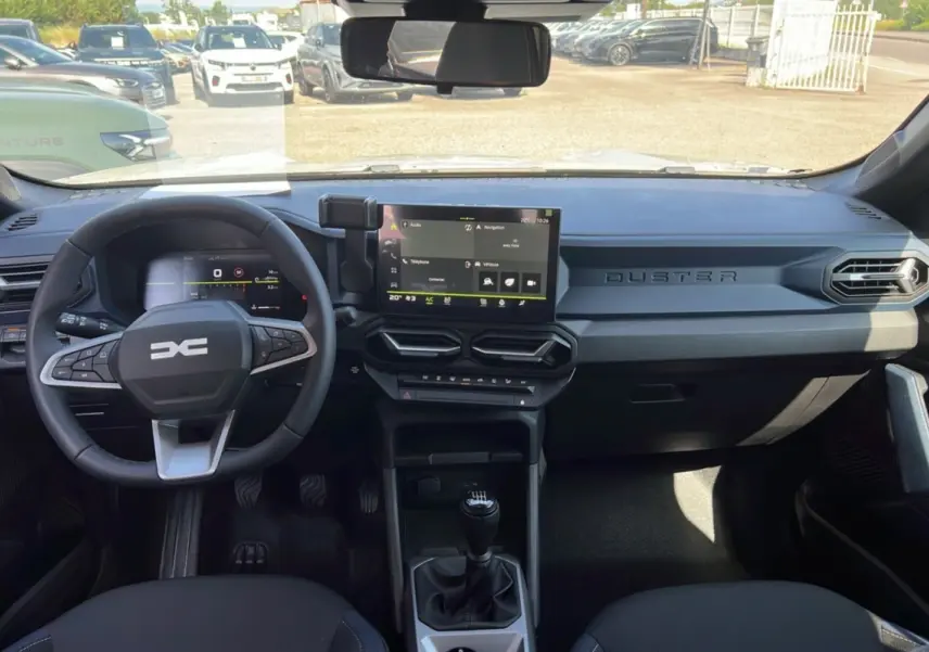 Vue intérieure du tableau de bord du Dacia Duster 2025 avec volant multifonction, écran tactile central et levier de vitesses manuel.
