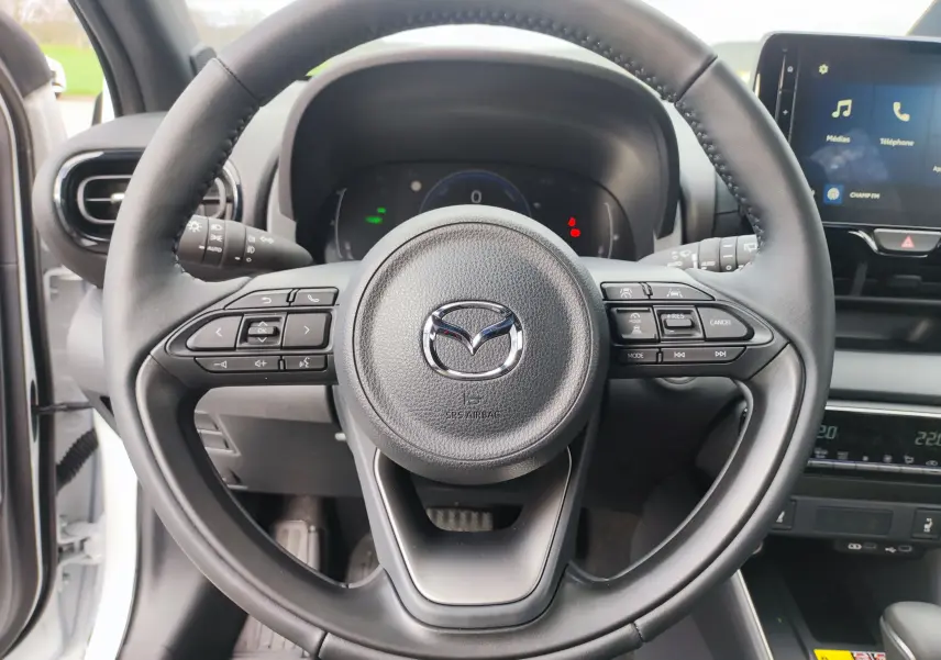 Vue rapprochée du volant noir de la Mazda2 Hybrid 2024, avec commandes intégrées et tableau de bord numérique visible.